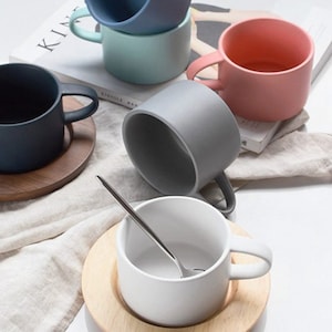 May include: A set of five ceramic mugs in various colors, including blue, green, gray, pink, and white. Each mug has a simple, modern design and is paired with a wooden saucer.