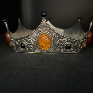 Könnte beinhalten: Eine Krone aus silberfarbenem Metall mit einem ovalen, bernsteinfarbenen Cabochon in der Mitte, flankiert von kleineren schwarzen runden Steinen. Das Band ist mit komplexen Prägemustern verziert und an jeder Spitze mit kleinen schwarzen Kugeln versehen.
