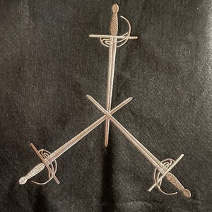 May include: Three silver swords embroidered on a black fabric background. The swords are arranged in a triangular shape with the points of the swords meeting in the center.