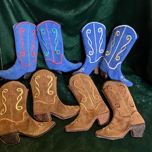 Puede incluir: Seis adornos de botas de vaquero bordadas, tres azules y tres marrones. Las botas azules tienen bordados blancos, rojos y verdes. Las botas marrones tienen bordados dorados.
