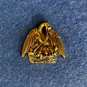 May include: A gold-colored metal pin featuring a pelican with outstretched wings, perched above a nest. The pin has intricate detailing and a vintage aesthetic. The pelican is the central focus, with its beak pointing downwards.