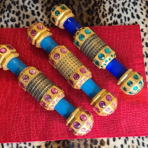 Può includere: Tre oggetti decorativi, di colore dorato, cilindrici con pietre preziose blu e viola. Ogni oggetto ha un diverso schema di pietre preziose e numeri.