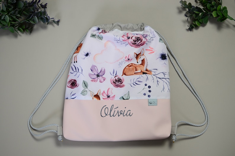 May include: Drawstring backpack with a watercolor floral and fawn design. The top portion features a white background with purple flowers, a fawn, and a cloud. The bottom is light pink with the name "Olivia" in script.