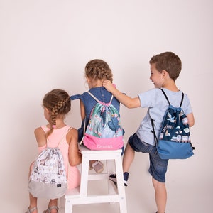 May include: Three children with drawstring backpacks. One backpack has a forest scene, another has a cactus design, and the third has a train and boat design. The cactus backpack has the name "Emma" printed on it. The backpacks are in various colors.