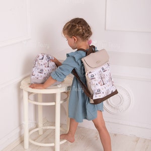 May include: A beige and brown backpack with a flap closure and a matching drawstring bag. Both bags feature a whimsical forest print with deer and trees. The backpack has brown straps and trim.
