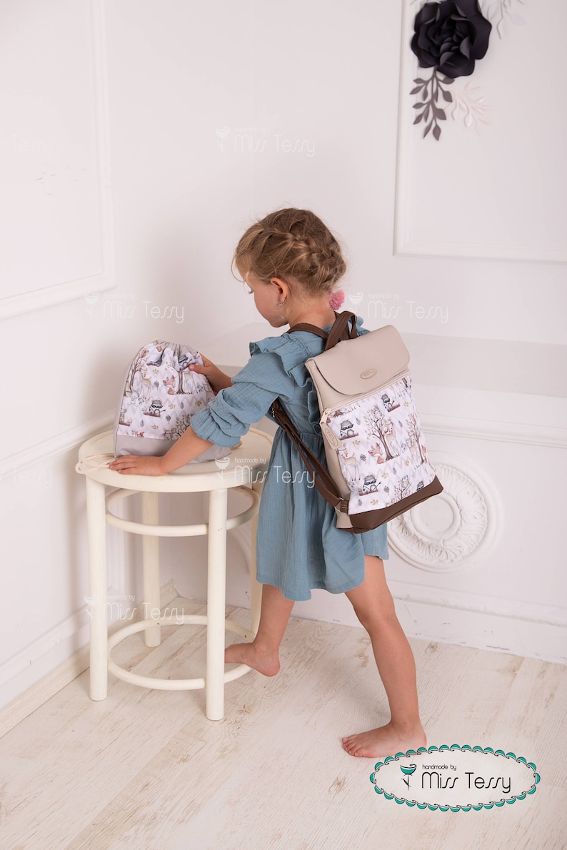 May include: A beige and brown backpack with a flap closure and a matching drawstring bag. Both bags feature a whimsical forest print with deer and trees. The backpack has brown straps and trim.