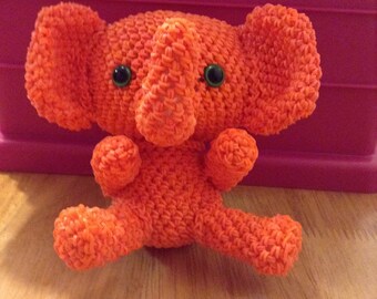 May include: An orange handmade elephant toy with large ears, a trunk, and small arms and legs. The elephant has black button eyes and is made from woven rubber bands. The toy is sitting on a wooden surface.