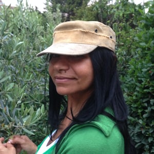 May include: A tan-colored, military-style cap with a short brim. The cap has a distressed look with visible stitching and small black rivets. The person in the image is wearing a green top and has long black hair.