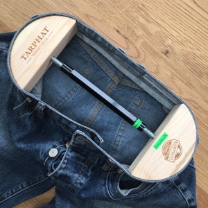 Può includere: Uno strumento in legno per allungare i jeans in denim, con una barra di metallo e due pezzi di legno con il testo "TARPHAT - TARPHAT.CO.UK" e "TARPHAT - TARPHAT.CO.UK" stampato su di essi.