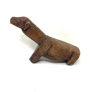 Könnte beinhalten: Eine braune, geschnitzte Holzfigur eines Hundes in sitzender Position. Der Hund hat geätzte Details am Körper und Kopf. Die Figur hat eine glatte Oberfläche und steht vor einem weißen Hintergrund. Die Ohren des Hundes sind sichtbar.