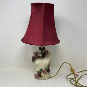 May include: A decorative table lamp with a burgundy shade and a mannequin-shaped base. The base is cream-colored and adorned with floral accents in shades of burgundy, cream, and green. The lamp has a cord with a plug.