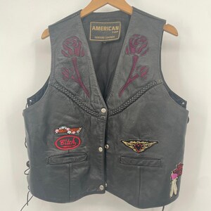 May include: Black leather vest with embroidered rose designs and patches. The vest features a button-up front, side laces, and a label that reads "AMERICAN TOP GENUINE LEATHER". Patches include the word "Bitch" and a Harley-Davidson logo.