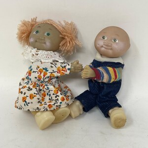 Könnte beinhalten: Zwei kleine Puppen mit Vinylköpfen und weichen Körpern. Eine Puppe hat orangefarbenes Garnhaar, einen weißen Spitzenkragen und ein cremefarbenes Kleid mit orangefarbenem Blumenmuster. Die andere Puppe ist kahl und trägt ein gestreiftes Oberteil unter einer dunkelblauen Latzhose.