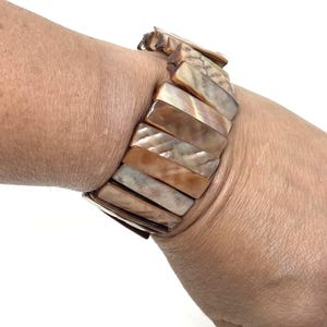May include: A stretch bracelet featuring rectangular, iridescent shell segments in shades of brown, tan, and cream. The bracelet is worn on a wrist, showcasing the natural variations in color and texture of the shell.