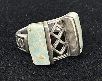 Puede incluir: Un anillo de plata con dos piedras rectangulares de color verde claro. El anillo tiene un diseño geométrico con un patrón de celosía entre las piedras. La banda presenta un patrón decorativo. El anillo está sobre un fondo negro.