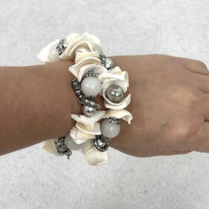 Puede incluir: Un brazalete con conchas blancas, cuentas grises y detalles metálicos plateados. El brazalete está compuesto por múltiples hilos, con las conchas dispuestas en un patrón decorativo. Las cuentas son redondas y lisas.