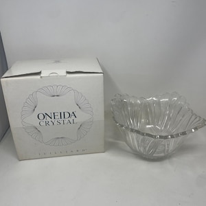 Könnte beinhalten: Eine klare Glasschale mit einem gewellten, blütenblattartigen Rand und vertikalen Rillen. Daneben steht ein weißer Karton mit der Aufschrift Oneida Crystal Juilliard, der einen kreisförmigen Zierrand und den Markennamen in dunkelblauem Text zeigt.