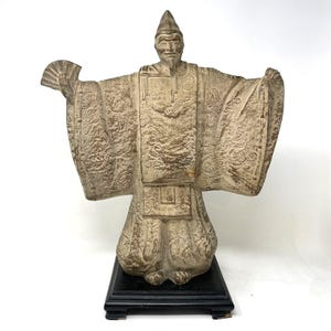 Op de afbeelding: Beige beeld van een man in traditionele Japanse kleding, met een waaier. Het beeld heeft gedetailleerde patronen op de kleding en staat op een zwarte rechthoekige basis. De armen van de figuur zijn uitgestrekt.