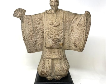 Op de afbeelding: Beige beeld van een man in traditionele Japanse kleding, met een waaier. Het beeld heeft gedetailleerde patronen op de kleding en staat op een zwarte rechthoekige basis. De armen van de figuur zijn uitgestrekt.