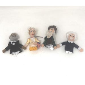 May include: Four finger puppets depicting historical figures. The puppets include a figure in a black suit, a woman in a yellow dress holding a flag, a man with a long white beard, and a man in a black suit with white hair.