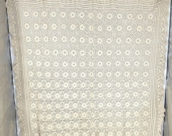 Könnte beinhalten: Cremefarbene, gehäkelte Decke mit floralem Muster. Die Decke hat einen gewellten Rand und eine detaillierte Bordüre. Das aufwendige Design zeigt ein sich wiederholendes Muster aus Blumen und durchbrochenen Quadraten, wodurch eine strukturierte Oberfläche entsteht.