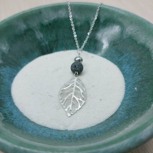 Peut inclure: Un collier en chaîne argentée avec un pendentif en forme de feuille délicate et une perle de lave noire. Le collier est présenté dans un bol vert et blanc.