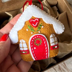 May include: A felt gingerbread house ornament featuring a brown body, a white roof, and a red door. The house is decorated with a red heart, green embroidered foliage, and two white windows with red accents. A red ribbon loop is attached to the top.