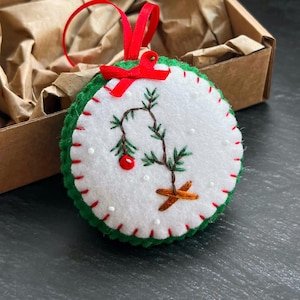 Peut inclure: Ornement de Noël fait à la main, rond, avec une base en feutre blanc, une bordure verte et des coutures rouges. Il présente un motif de sapin de Noël brodé avec une baie rouge et un ruban rouge pour l'accrocher.