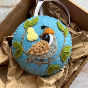 Könnte beinhalten: Ein rundes, hellblaues Filzornament mit einer handgestickten Wachtel mit braun-grauem Körper, beigem Kamm und einer gelben Birne. Das Ornament ist mit grünen Filzblättern an braunen gestickten Zweigen verziert und hat eine weiße Bandschlaufe.
