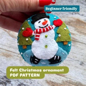 May include: A round felt Christmas ornament featuring a white snowman wearing a black hat, red scarf, and red mittens, ice skating on a blue background with two green pine trees and white beads. The text reads Beginner friendly and Felt Christmas ornament PDF PATTERN.