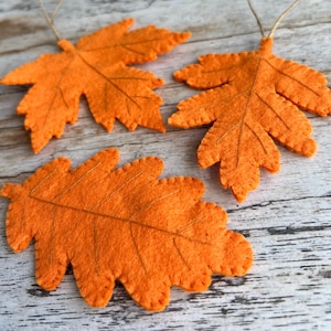 Könnte beinhalten: Drei orangefarbene Filzblatt-Ornamente mit braunen Nähten und Schnuraufhängern. Die Blätter sind auf einer hellen Holzmaserung angeordnet. Die Blätter sind etwa 15 cm lang und 10 cm breit.