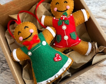 May include: Two felt gingerbread person ornaments with red hanging ribbons. One wears a green dress with a red heart and white trim, the other a red outfit with green pockets, white trim, and a holly sprig. Both have stitched smiles and pink cheeks.