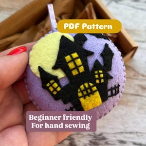 May include: A circular, purple felt decoration featuring a black haunted house design, a yellow moon, and yellow window details. It has a white ribbon for hanging and the text "PDF Pattern" and "Beginner friendly For hand sewing".