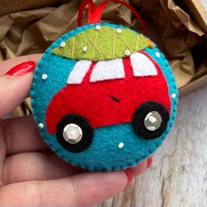 May include: A handmade felt Christmas ornament depicting a red car with a green tree on its roof, set against a blue backdrop. The ornament has white details, black wheels with silver sequins, and a red hanging ribbon.