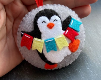 Può includere: Un ornamento di pinguino in feltro con corpo bianco, ali nere e becco arancione. Il pinguino tiene una stringa di bandierine di feltro colorate in rosso, giallo, blu e verde.