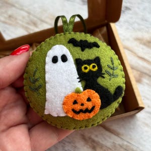 May include: A circular, green felt Halloween decoration with a white ghost, black cat, and carved pumpkin. A black bat and green leaves are also stitched on the ornament. A green ribbon is attached for hanging.