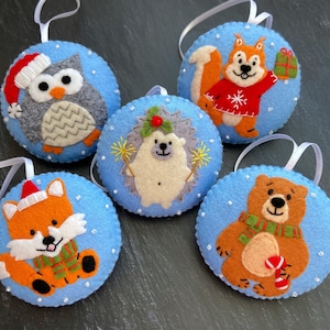 May include: Five felt ornaments with blue backgrounds and white polka dots. Each ornament features a different animal: an owl, a hedgehog, a squirrel, a fox, and a bear. The animals are all wearing festive attire.