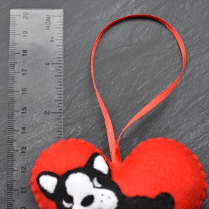 May include: Red felt heart-shaped ornament with a black and white felt French bulldog design. The ornament is attached to a red ribbon.