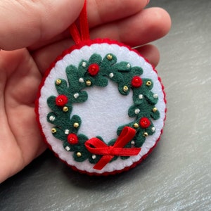 K&ouml;nnte beinhalten: Ein Weihnachtsbaumschmuck aus Filz in Form eines Kranzes mit rotem und gr&uuml;nem Filz und goldenen und wei&szlig;en Perlen. Der Kranz hat eine rote Schleife aus Filz.