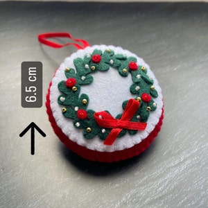 K&ouml;nnte beinhalten: Ein Weihnachtsbaumschmuck aus Filz in Form eines Kranzes mit roten Beeren, goldenen Perlen und einer roten Schleife. Der Kranz hat einen Durchmesser von 6,5 cm.