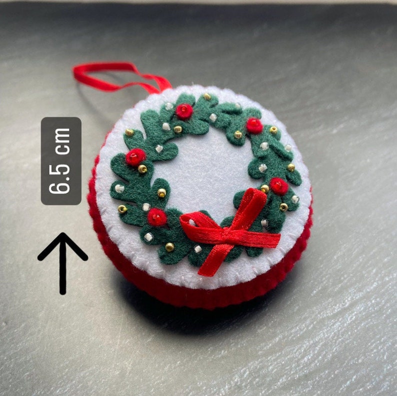 K&ouml;nnte beinhalten: Ein Weihnachtsbaumschmuck aus Filz in Form eines Kranzes mit roten Beeren, goldenen Perlen und einer roten Schleife. Der Kranz hat einen Durchmesser von 6,5 cm.