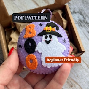 May include: A round purple felt ornament featuring a white ghost wearing a black witch hat with an orange band. The text "PDF PATTERN" appears at the top, and "Beginner friendly" is on the right. The word "BOO" is stitched in orange and black.