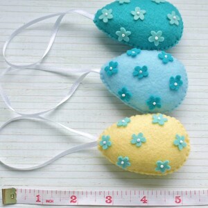 May include: Three felt Easter eggs with white ribbon hangers. The eggs are decorated with small felt flowers in shades of blue and yellow.