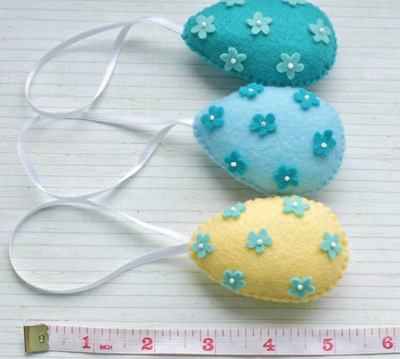 May include: Three felt Easter eggs with white ribbon hangers. The eggs are decorated with small felt flowers in shades of blue and yellow.