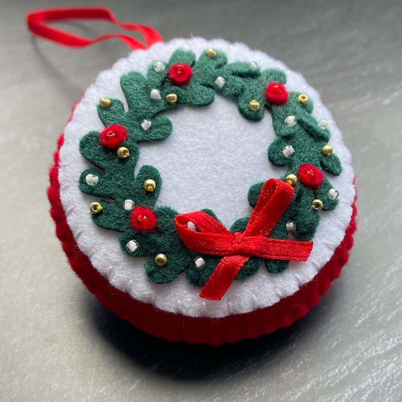 K&ouml;nnte beinhalten: Ein Filz-Weihnachtsornament in Form eines Kranzes mit roten und wei&szlig;en Perlen, gr&uuml;nen Bl&auml;ttern und einer roten Schleife.