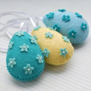 May include: Three felt Easter eggs decorated with blue and yellow flowers. The eggs are hanging from white ribbon.