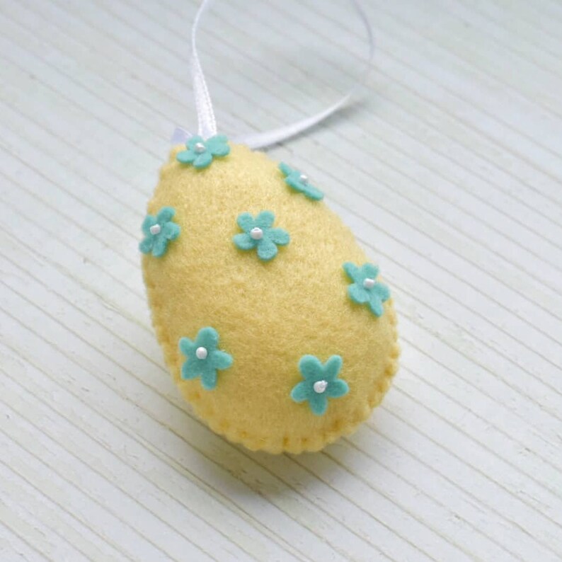 May include: A yellow felt Easter egg ornament with a white ribbon and blue felt flowers. The flowers have white centers.