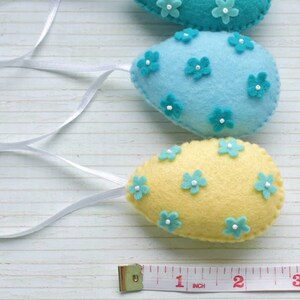 May include: Three felt Easter eggs with blue and yellow fabric and blue flower embellishments. The eggs are strung together with white ribbon and a measuring tape is in the bottom of the image.