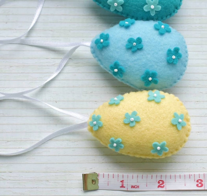 May include: Three felt Easter eggs with blue and yellow fabric and blue flower embellishments. The eggs are strung together with white ribbon and a measuring tape is in the bottom of the image.