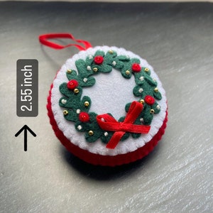K&ouml;nnte beinhalten: Ein Weihnachtsbaumschmuck aus Filz in Form eines Kranzes mit einer roten Schleife. Der Kranz ist mit gr&uuml;nen Bl&auml;ttern, roten Beeren und goldenen Perlen verziert. Der Schmuck ist 2,55 Zoll im Durchmesser.
